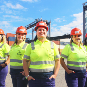 Five women wearing safety gear stand in front of container cranes and stacked shipping containers at DP World’s Santos terminal in Brazil.