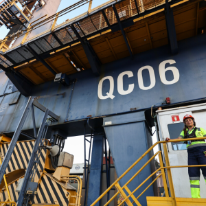 Female port worker wearing a hard hat stands on a platform beside a large quay crane labeled QC06 at DP World’s Santos terminal in Brazil.