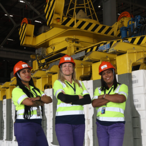 Three women wearing high-visibility uniforms and hard hats stand in front of large cargo-handling machinery inside a DP World warehouse facility in Brazil.