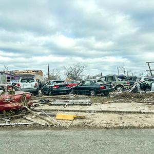 A group of damaged cars and building, debris has broken windows and is strewn around the street and lot.