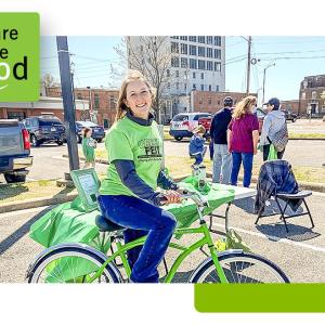 Shelly Gist on a green bicycle next to a table outdoors.