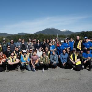 A large group of DP World employees and community volunteers posing together near the coastline with mountains and water in the background during a cleanup event.