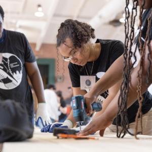 Person using a tool with another person helping and someone watching at the side