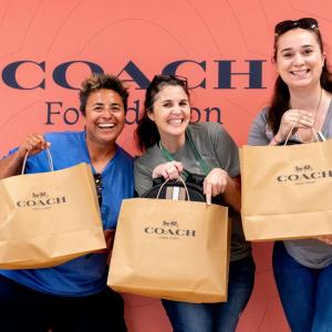 Three woman stand together holding Coach shopping bags with in front of a salmon pink colored backdrop with the Coach logo