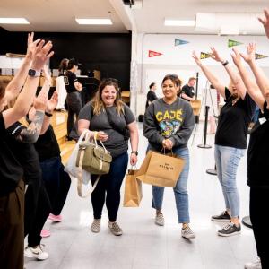 People standing in two lines with their arms raised while two women holding Coach shopping bags walk down the middle
