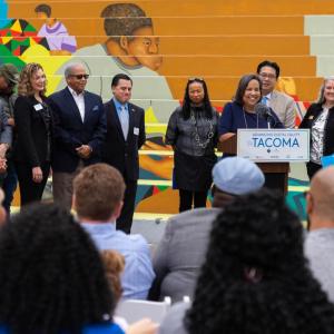 Mayor Victoria Woodards at a podium, others behind them and bleachers in the background. Audience seated in front.
