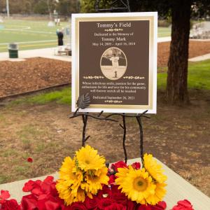 A commemorative plaque for Tommy Mark is positioned by "Tommy's Field" surrounded by flowers. 