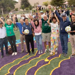 Members of the Mark Family, community members and representatives of the LA Galaxy and LAFC celebrate with a cheer and their hands in the air in the middle of "Tommy's Field." 