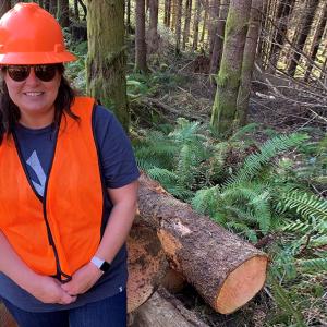 Tracie in front of cut timber in a forest wearing a safety hat and vest.