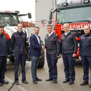 Six people stand in a line, the middle two shake hands. A fire truck and tractor behind them.