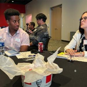 Truman and another talking at a table in a room with others. KFC bucket central.