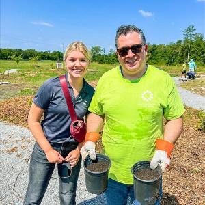 Two people stood next to each other smiling, the person on the right is holding two plant pots