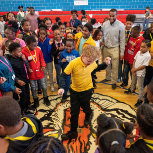 a large group of kids in a gymnasium, circled around one who is dancing in the middle.