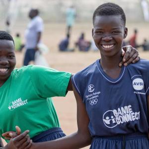 Two athletes smiling, holding hands, one with their arm over the other. A sandy field behind them and others in the distance sitting.