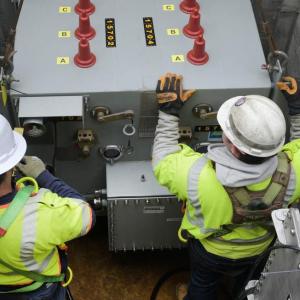 Two people in protective uniform working on a large electrical unit underground.