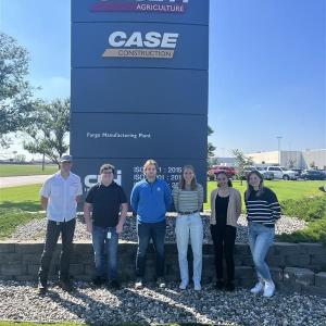 Six people posed outside in front of a tall sign for Case.