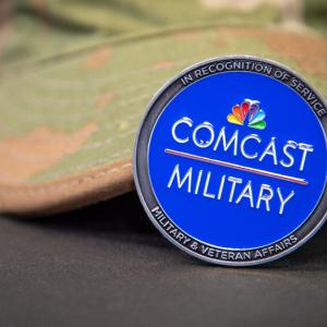 Pin with Comcast Military on it. A camouflage hat behind it. 