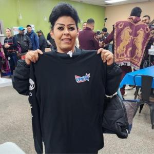 a person holding up a black long-sleeve shirt with VPP on the front pocket. Others in the room walking around.