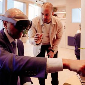 A seated person in a suit using a VR headset, pointing off screen. A person standing next to them talking.