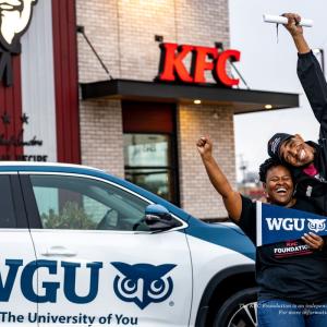 Two people smiling with one arm raised. One holds a WGU pendant. Standing in front of a car with WGU and logo on the side and all in front of a KFC restaurant.