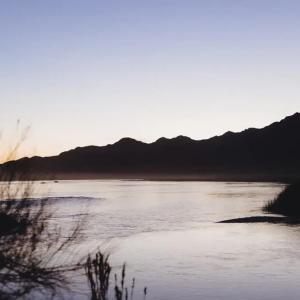 Landscape of a waterway, tall grass, and hilly terrain. A setting sun to the side.
