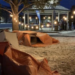 Makeshift shelters outside on a snowy ground. People gathered by a gazebo with string lights.