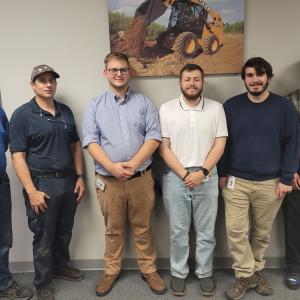 Six people posed in a hallway in front of an image of a case front-loader.
