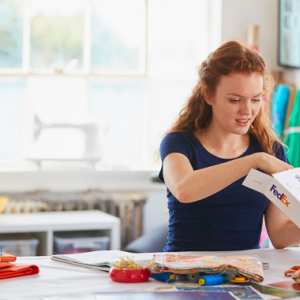 a person in a bright room, sitting at a large table with craft items, opening a FedEx box