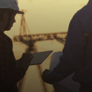 Two professionals wearing safety hats looking at a tablet and piece of paper