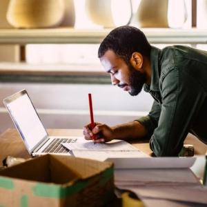 A person leant on a desk writing in a book that it resting on a laptop