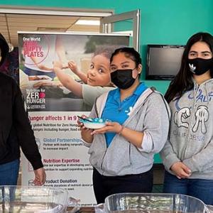 three people standing in a library next to a "One world zero empty plates" poster, one holds a bowl.