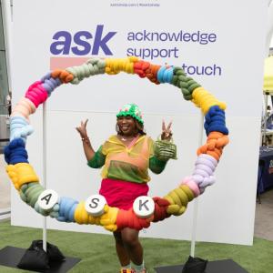 woman holding up peace signs in a giant friendship bracelet with the word "ASK"