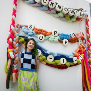 woman standing in front of giant friendship bracelets with the words "acknowledge" and "support"