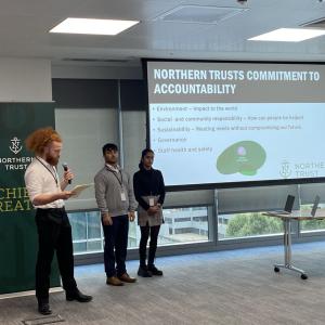 Three people standing at the front of the room, one speaking in to a microphone. A projected dispay to the side.