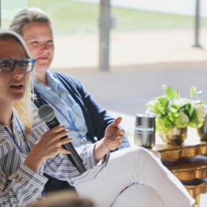  A person with a microphone, seated, others to their side in a sunny indoor room.
