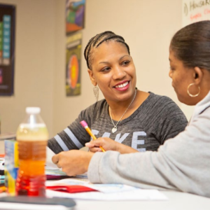 Two people sitting at a desk, one holding a pencil, posters and a periodic table on the walls.