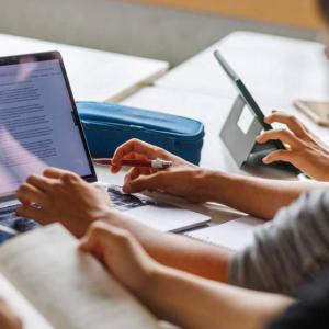 Looking over the shoulder of a person with an open book, another to their right using a laptop on a desk.