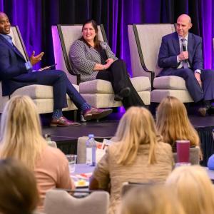 Three people seated on a stage, talking to a seated audience.