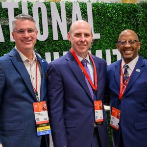 Three people posed in front of a hedge with "National Alice Sumit" sign.