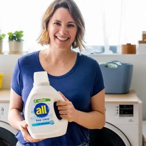 A smiling person holding a bottle of all detergent in front of a washer and dryer.