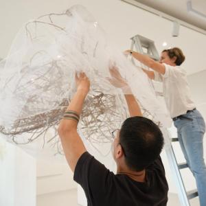 two people attach a gauzy textile sculpture to a gallery ceiling
