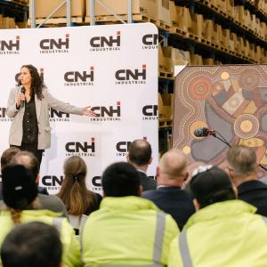 A person presenting next to artwork in front of a crowd, some in safety clothing. all in a warehouse.