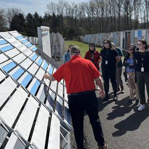 A person speaking to a group outside, a rack of painted boards to the side.
