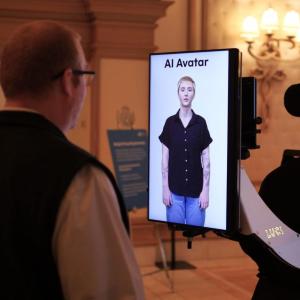 Erin Taylor appearing as an avatar on a small screen, another looks at it.