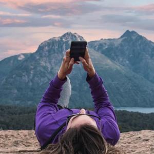 A person laying on a rock looking at their phone held above them. A mountain landscape in the distance.