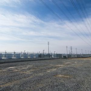  A power station with tall power lines above and graveled pad in front.