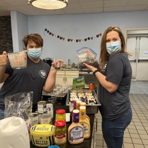 Two Bath & Body Works associates stand at a counter in the Ronald McDonald House kitchen. One associate is holding up a measuring cup filled with yogurt and a small cup of blueberries. The other associate is holding up a bag of blueberries. On the counter are small cups filled with yogurt and topped with fresh fruit.