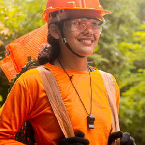 A smiling person in bright orange and safety gear in a forest setting.
