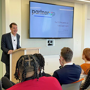 William Demchak standing at a podium speaking to a room of students and adults. A screen behind him displays "Partner up"