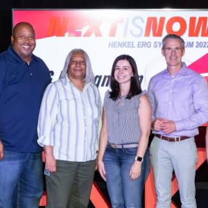 A group of people in a line posing for the camera. "Next it Now" banner behind them.